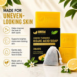 Three AMVital Natural lemon-turmeric kojic acid soap bars with a black box and a mesh soap bag, styled with lemon slices, white flowers, and green leaves on a light marble surface with text overlays on the left.
